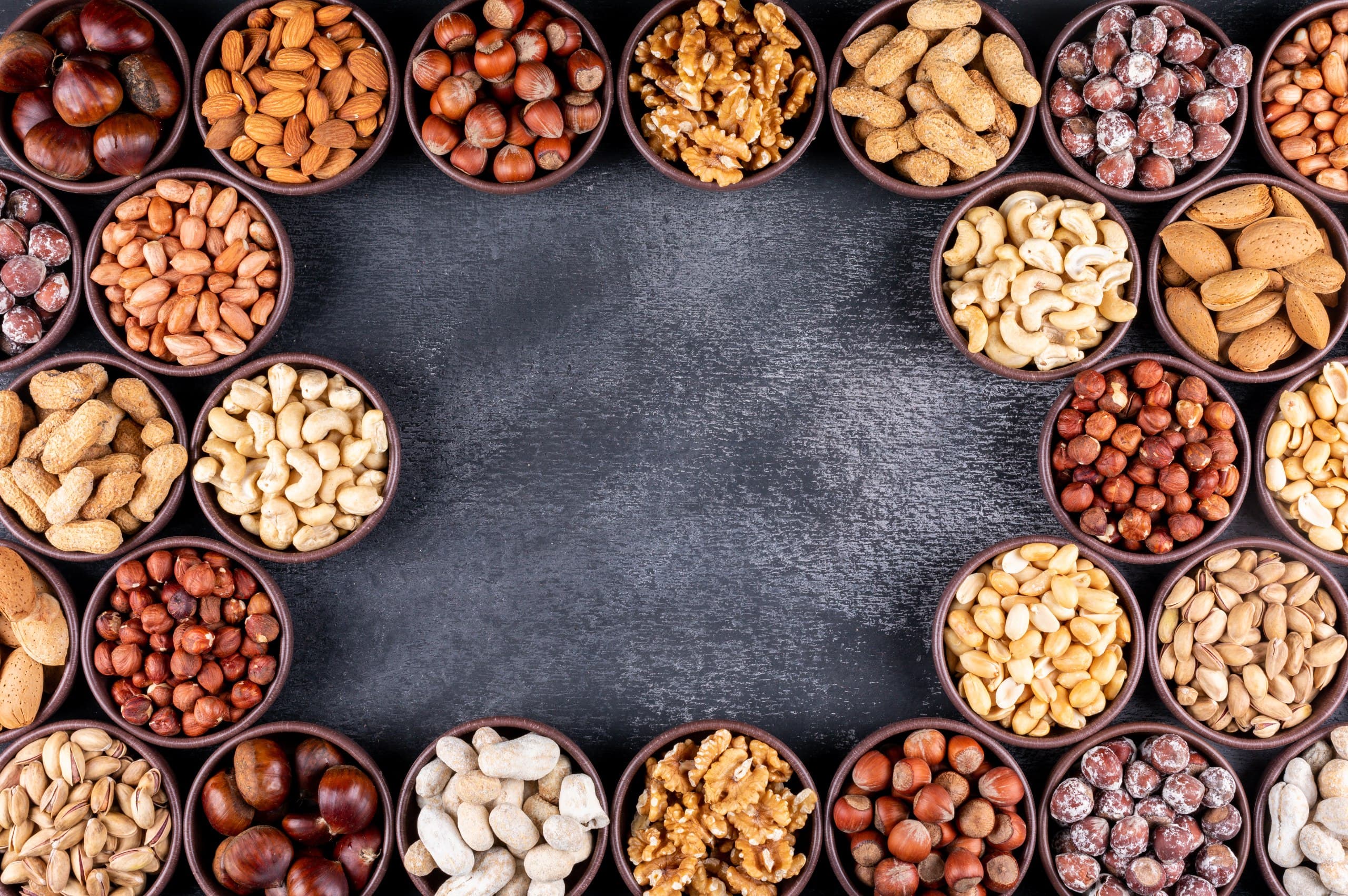 verschiedene nuesse darunter mandeln cashews walnuesse haselnuesse pistazien und erdnuesse in runden braunen schalen auf schwarzer schieferplatte von oben